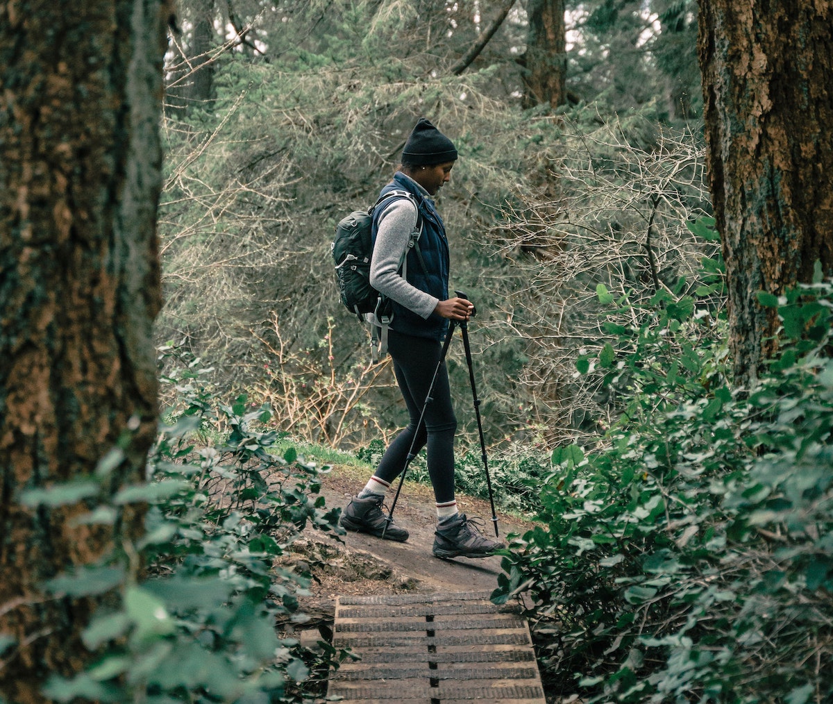 A woman of color hiking along a trail in the Pacific Northwest. She is surrounded by tall trees and lush vegetation.